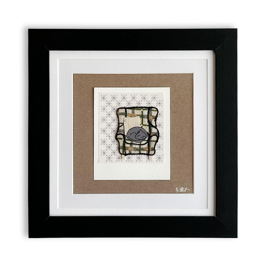 A framed art print featuring a stylized paper cut of a plaid armchair with a gray cat, set against a patterned cream background. The print is mounted on a brown paper mat and enclosed in a white and black frame.