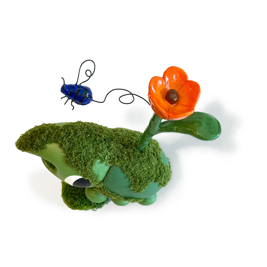 A whimsical green bug sculpture covered in faux moss, with a bright orange flower on a stem and a small blue wire beetle above it, photographed on a clean white background.