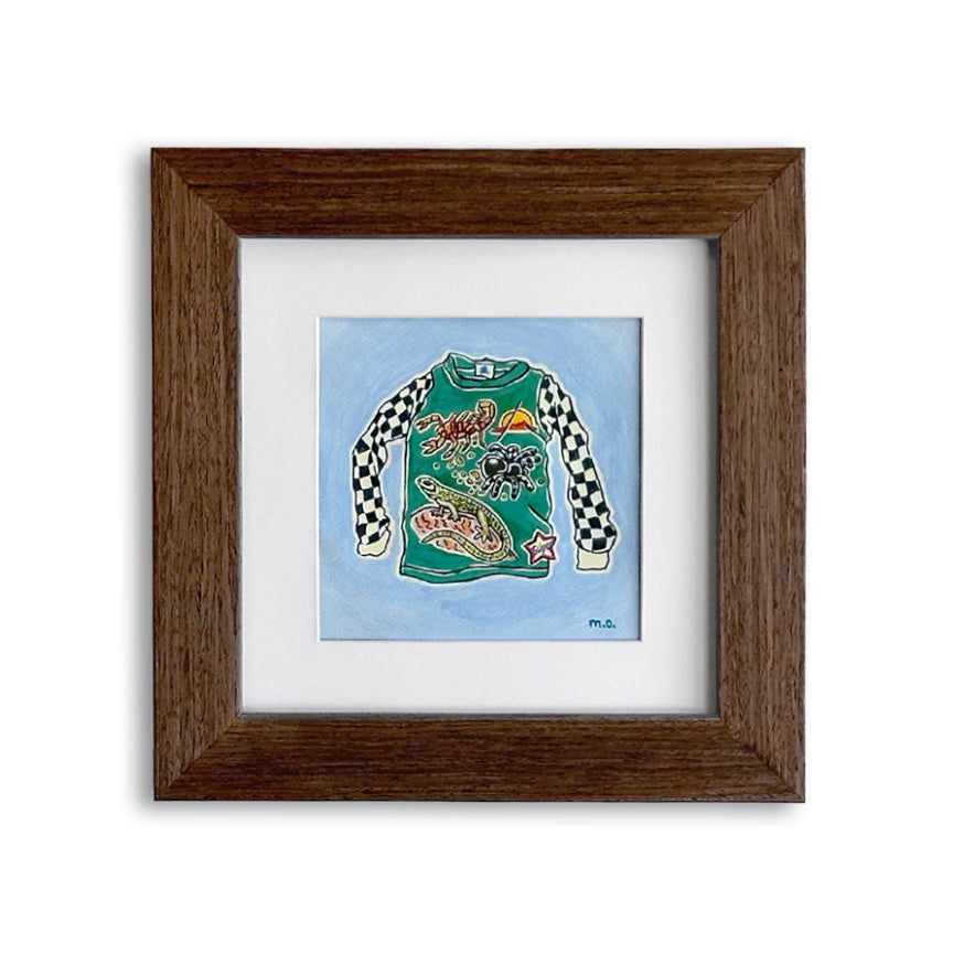 A framed art print featuring a colorful illustration of a long-sleeve motocross shirt covered in desert wildlife patches on a light blue background, set within a white mat and a dark brown wooden frame.