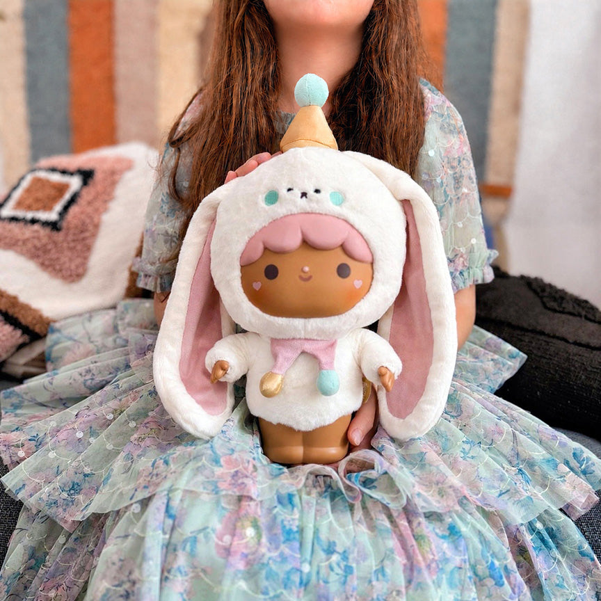 A child in a pastel floral dress holds the plush Momiji doll. The background shows a cozy indoor setting with pillows and a striped wall.