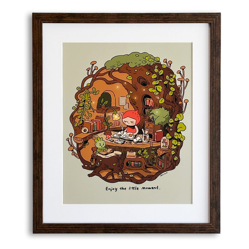 A framed art print featuring a whimsical illustration of a child with red hair sitting at a table and writing in a book inside a treehouse, surrounded by green vines and books. A little plant creature is sitting at a smaller desk next to her, reading a book. Under the scene are the words "Enjoy the little moment." The print is mounted with a wide white mat and enclosed in a dark brown wooden frame, set against a plain white background.