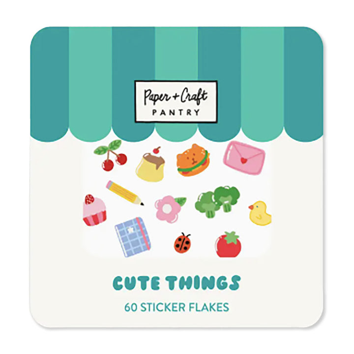 A package of Paper + Craft Pantry Trinkets sticker flakes. The packaging has a cute storefront design with blue and turquoise stripes, a white window displaying various cute character stickers, including a pink envelope, green broccoli, yellow duck, red strawberry, orange burger, blue notebook, pink flower, cherries, and other small motifs, and blue text at the bottom reading &