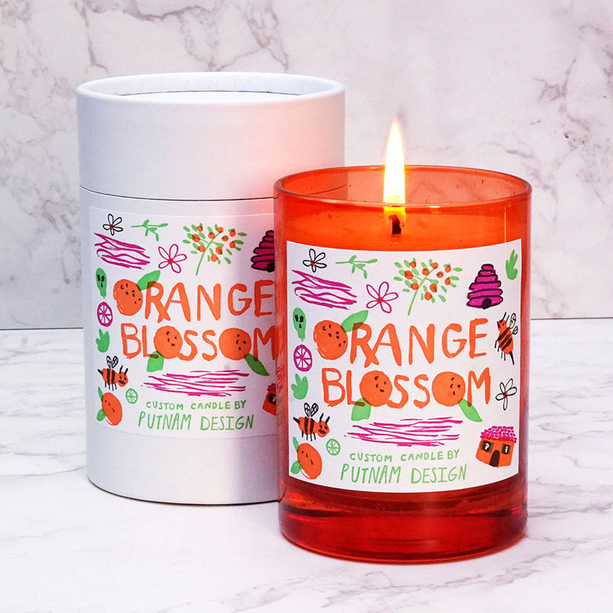 Orange glass candle with illustrated decorative label and the text "Orange Blossom custom candle by Putnam Design" on a marble background. 