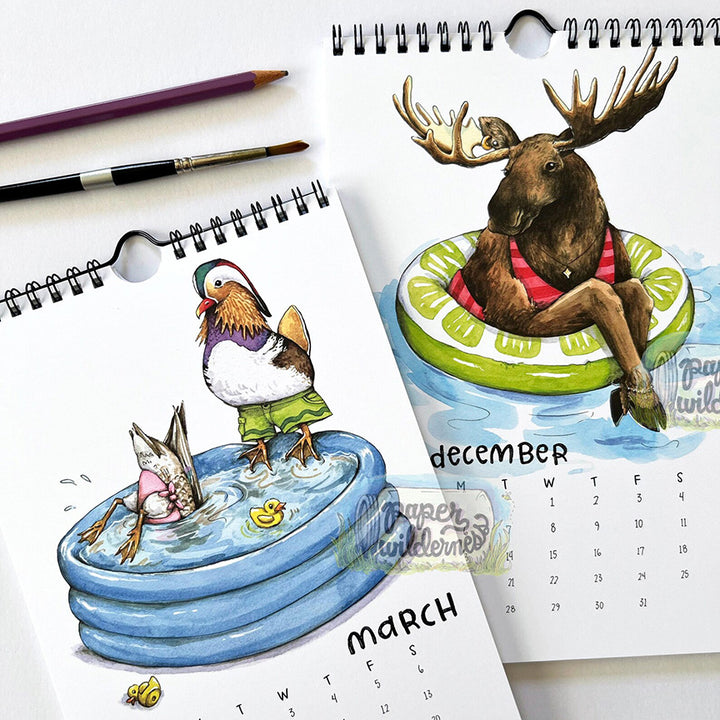 Two spiral-bound monthly calendar pages with watercolor illustrations of a duck wearing green swim shorts standing in a blue blow-up pool and a moose sitting in a lime-green float. 