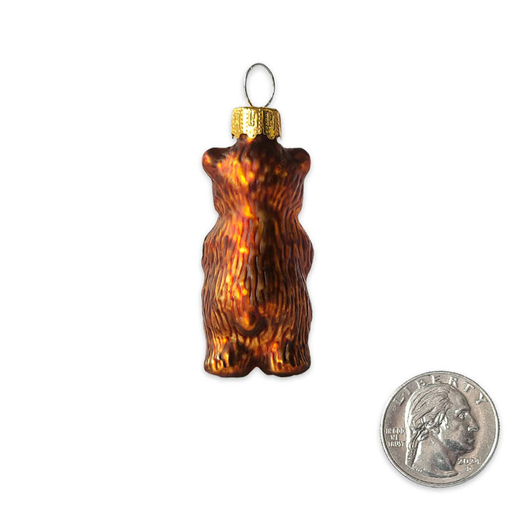 The backside of molded-glass ornament in the shape of a small brown bear, next to a quarter for size reference. Backside of bear is all brown and yellow with a little tail. The quarter is about half the size of the ornament. 