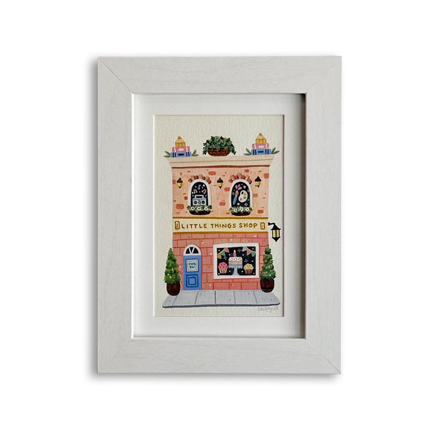 An art print featuring a whimsical illustration of a brick building labeled "Little Things Shop" on a yellow sign. A cake is displayed in the bottom window, and a paint palette and boom box shown in the top two windows. The piece is shown in a white frame against a white background.