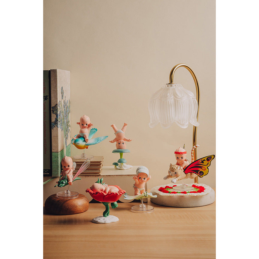 A collection of whimsical, pastel-colored fairy and cherub figurines displayed on a wooden tabletop next to a vintage book and a small gold-framed bell lamp against a warm beige wall.