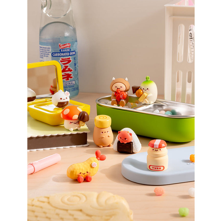 A styled tabletop scene with several small cute Smoko snack character figurines and toy accessories arranged on a light wood surface. 