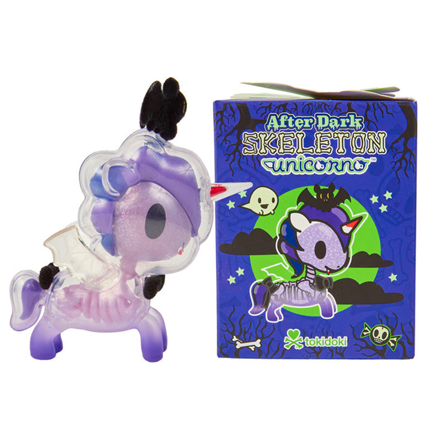 A Tokidoki After Dark Skeleton Unicorno collectible figure shown next to its blue illustrated packaging box on a plain white background. The Unicorno figure is clear so that you are able to see it's purple skeleton on the inside of the figure. It also has a bat on top of it's head and two black bows. 
