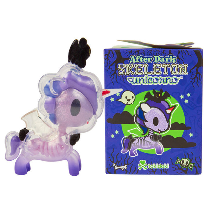 A Tokidoki After Dark Skeleton Unicorno collectible figure shown next to its blue illustrated packaging box on a plain white background. The Unicorno figure is clear so that you are able to see it&