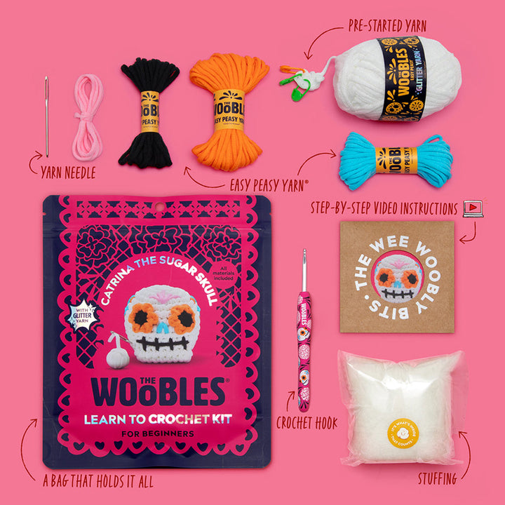 All materials included in Wooble kit (yarn needle, yarn, themed crochet hook, instructions, stuffing, and reusable zip bag) on a pink background. 