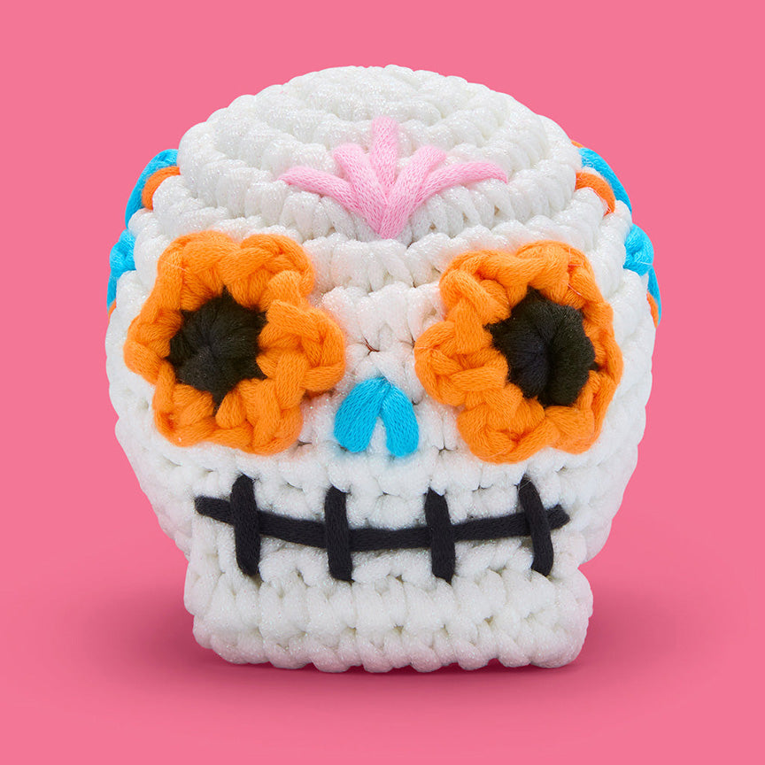 Finished crochet— a white sugar skull with orange petals surrounding in black eyes, black stitched mouth, blue skull nose, and light pink design on it&