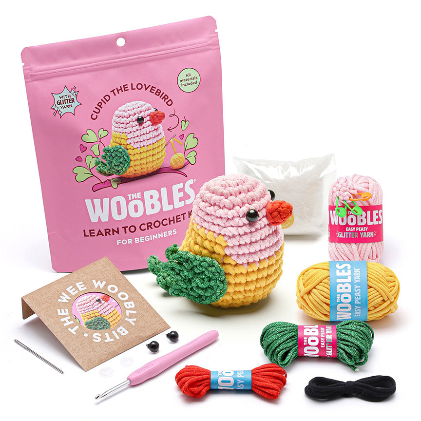 All materials included in Wooble kit (yarn needle, yarn, plastic eyes, pink crochet hook, instructions, stuffing, and reusable zip bag) on a white background. 
