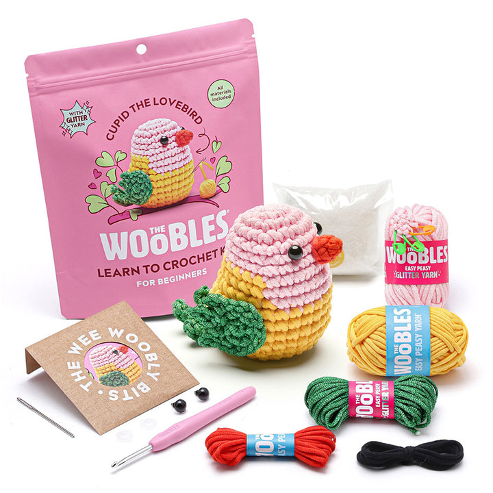 All materials included in Wooble kit (yarn needle, yarn, plastic eyes, pink crochet hook, instructions, stuffing, and reusable zip bag) on a white background. 