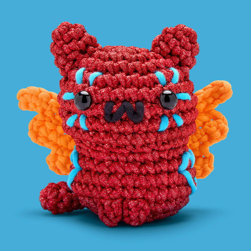 Finished crochet— a red cat with orange wings and blue stitch design surrounding it's eyes and on it's side. 