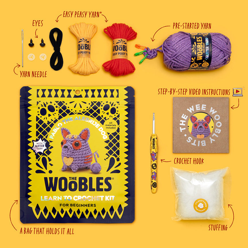 All materials included in Wooble kit (yarn needle, yarn, plastic eyes, themed crochet hook, instructions, stuffing, and reusable zip bag) on a yellow background. 
