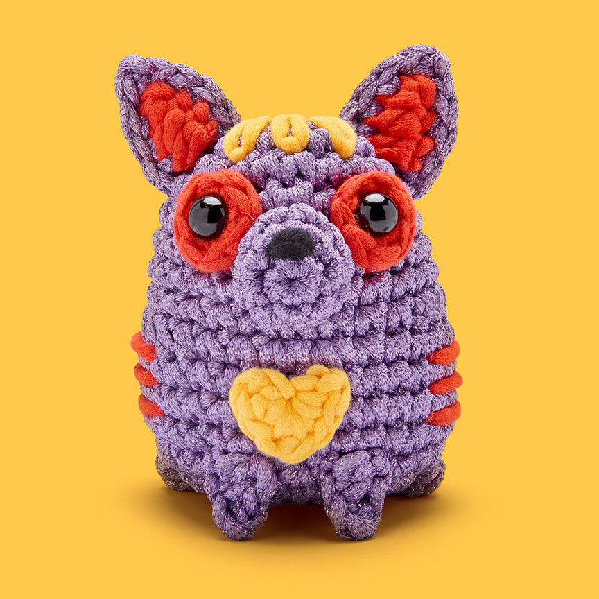 Finished crochet— a purple Chihuahua-looking dog with red circles around it's eyes, red inside ears, and red stripes, with a yellow pattern on it's head and a yellow heart patch on it's chest. 
