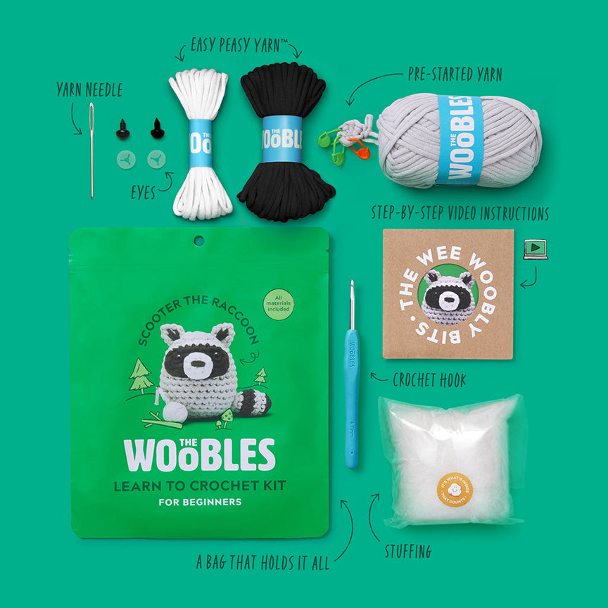 All materials included in Wooble kit (yarn needle, yarn, plastic eyes, blue crochet hook, instructions, stuffing, and reusable zip bag) on a green background. 