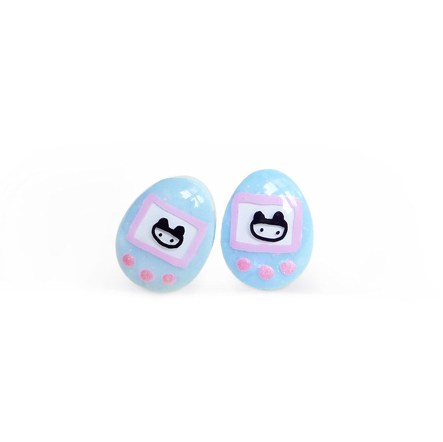 A pair of glittery blue stud earrings shaped like Tamagotchis, an egg-shaped mini-game console with a small pixel creature on its screen.